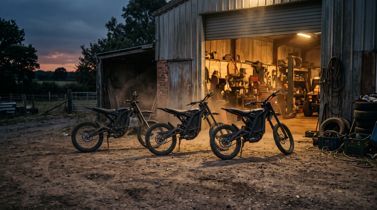 Three electric dirt bikes lined up on packed dirt outside a workshop at dusk